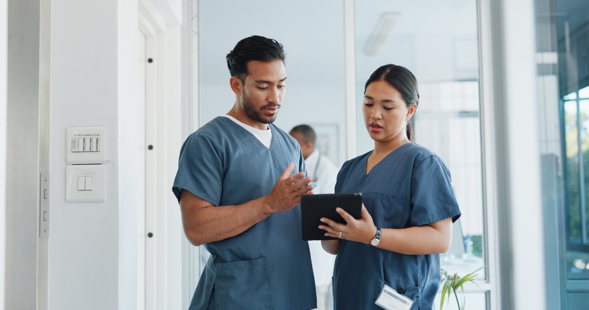 Two healthcare professionals in scrubs review workforce information on a tablet in a hospital corridor.