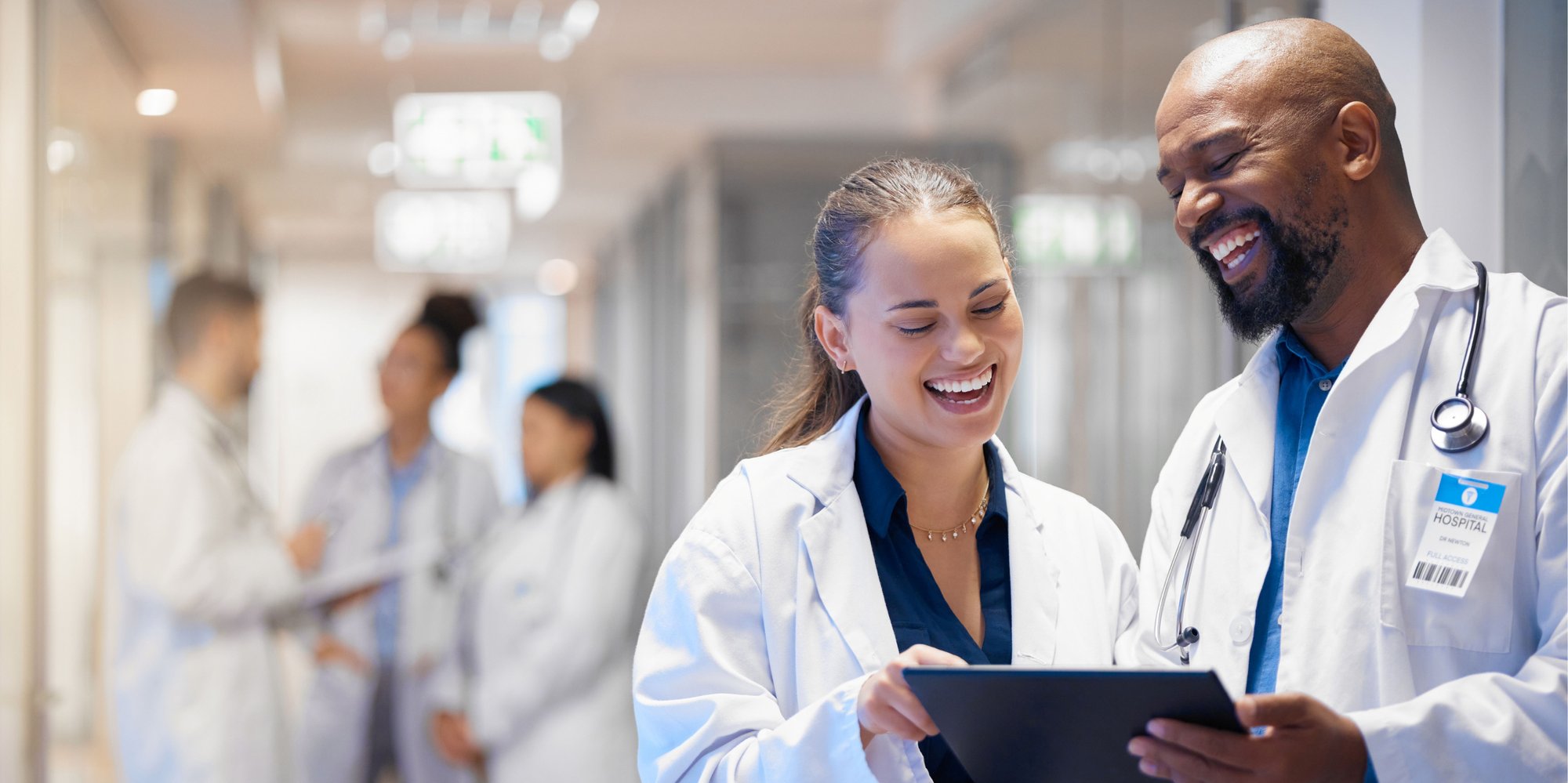 A female doctor and male doctor are standing in a hospital hallway smiling and looking at an iPad. Behind them three other clinicians are gathered around talking.