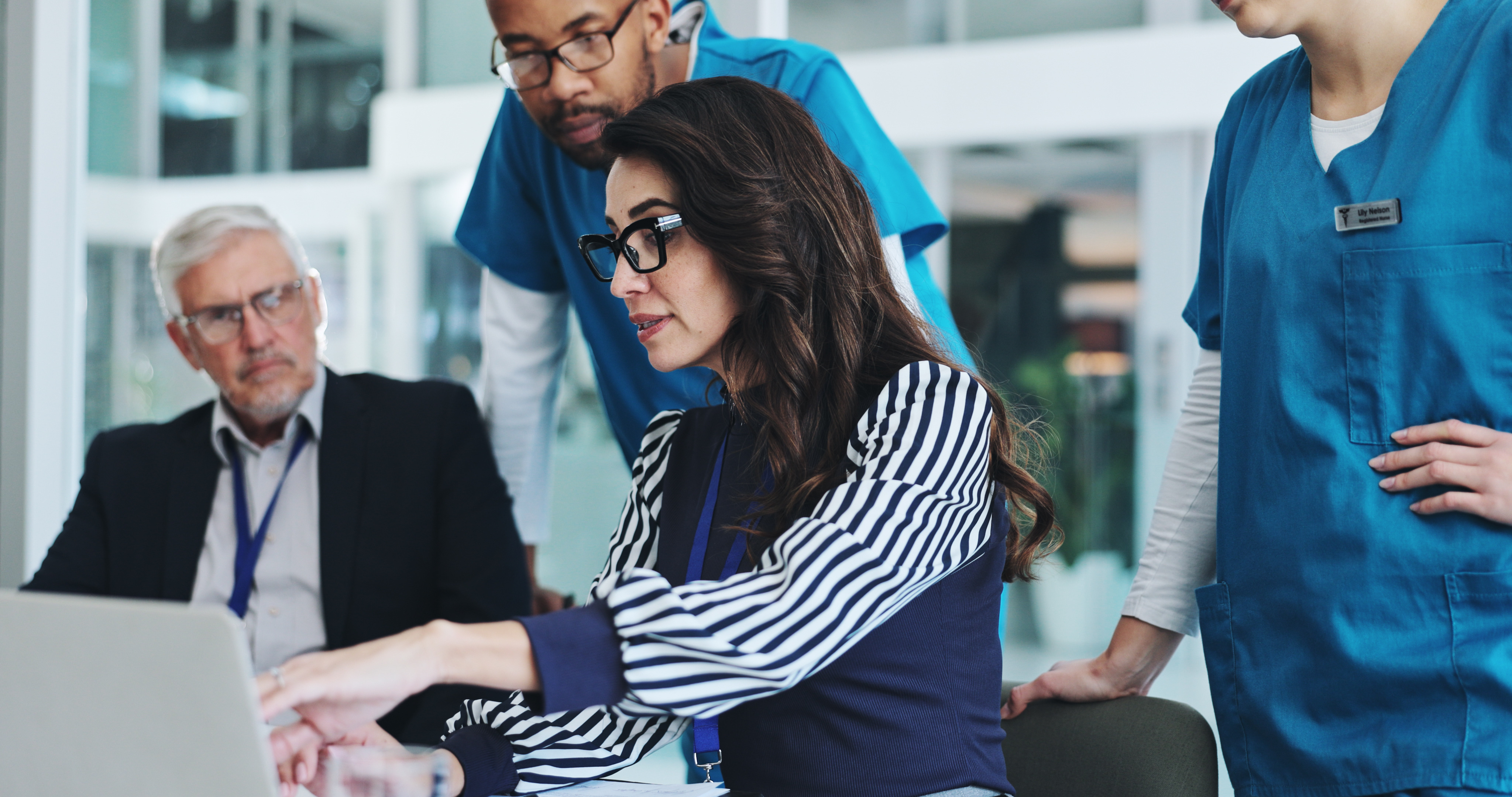A team of healthcare professionals and a business leader collaborate around a laptop in a modern office setting, reviewing information together.