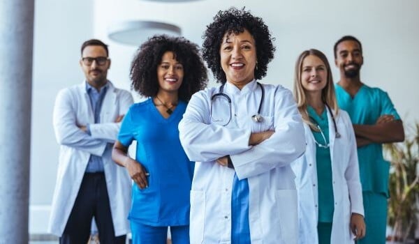 A diverse group of five healthcare professionals, two doctors in white coats and three clinicians in scrubs, stand together in a bright clinical setting, smiling confidently with arms crossed.
