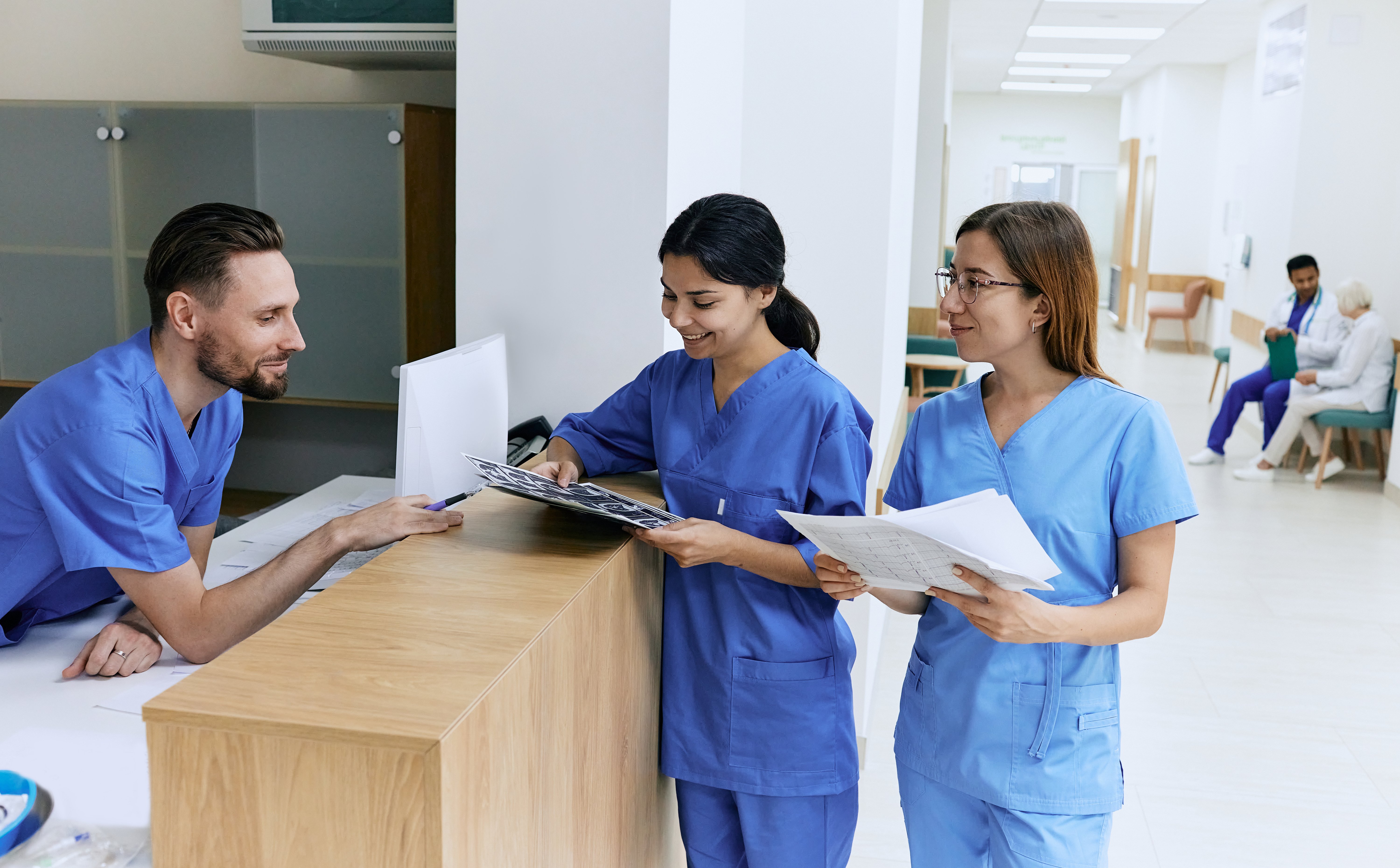 Three nurses are standing at a desk in the middle of a hospital corridor, smiling at each other. The male nurse behind the desk is standing. He has dark brown hair and a dark brown beard, wearing blue scrubs, and leaning forward on the desk. He is looking at MRI scans that the middle nurse is holding. The middle nurse is also in blue scrubs with dark hair that is in a ponytail. To her left is the third nurse who is wearing light blue scrubs. The third nurse has lighter brown hair and is wearing glasses. She is holding some papers. Over her left shoulder you can see an elderly patient sitting in a chair talking to a doctor. 
