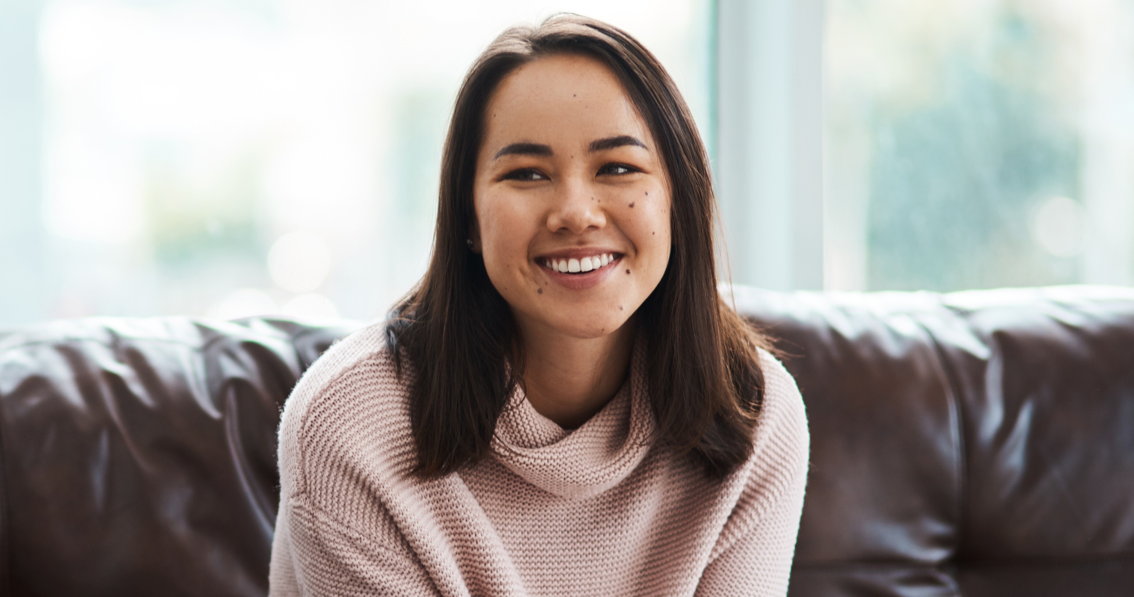 Smiling young woman sitting on a couch in a bright, modern behavioral health office setting, suggesting a calm and supportive mental health care environment.