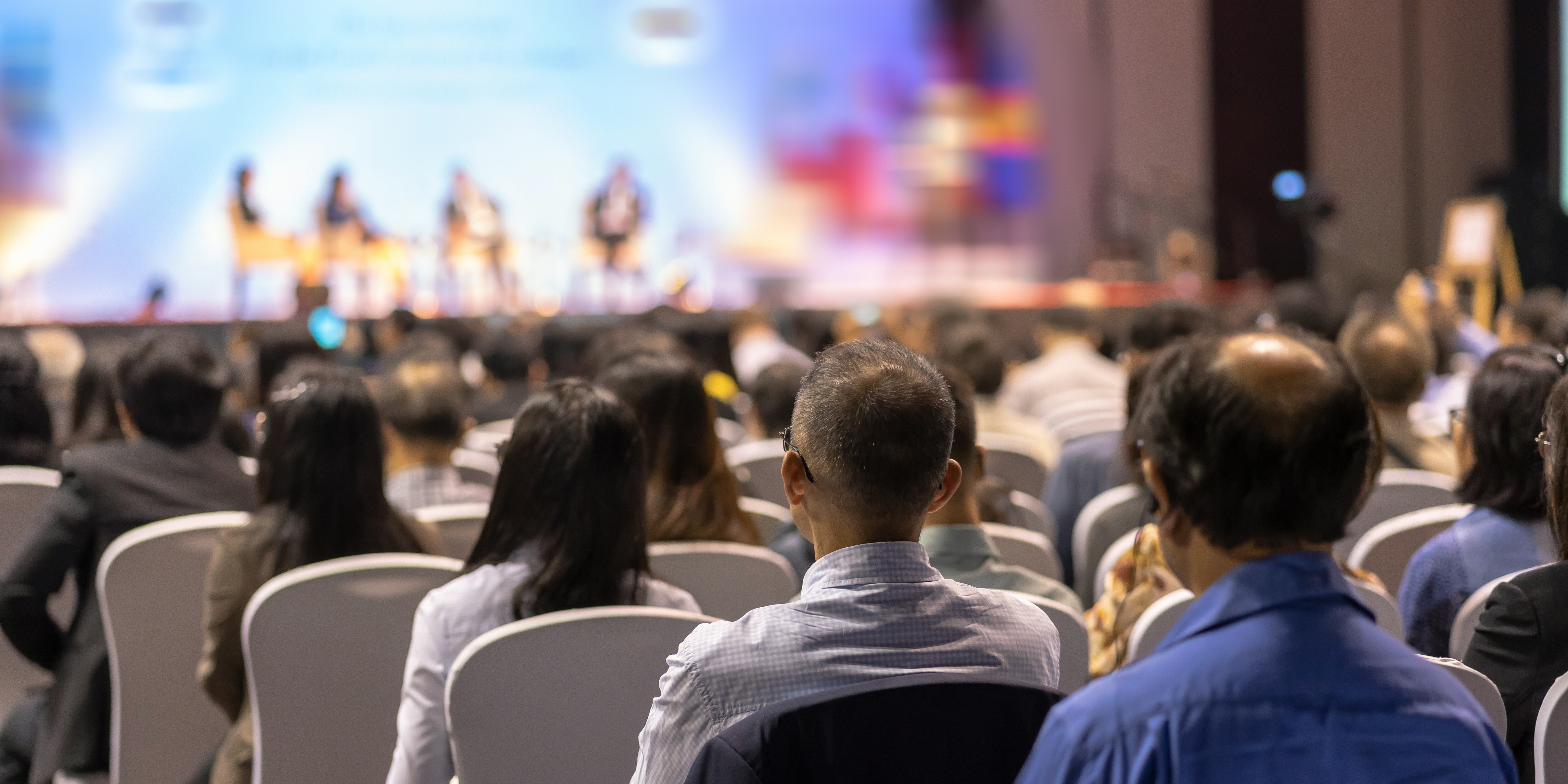 Photo from a conference speaking session. Image shows professionals sitting in chairs facing a stage. On stage a panel consisting of several speakers is sitting in chairs.