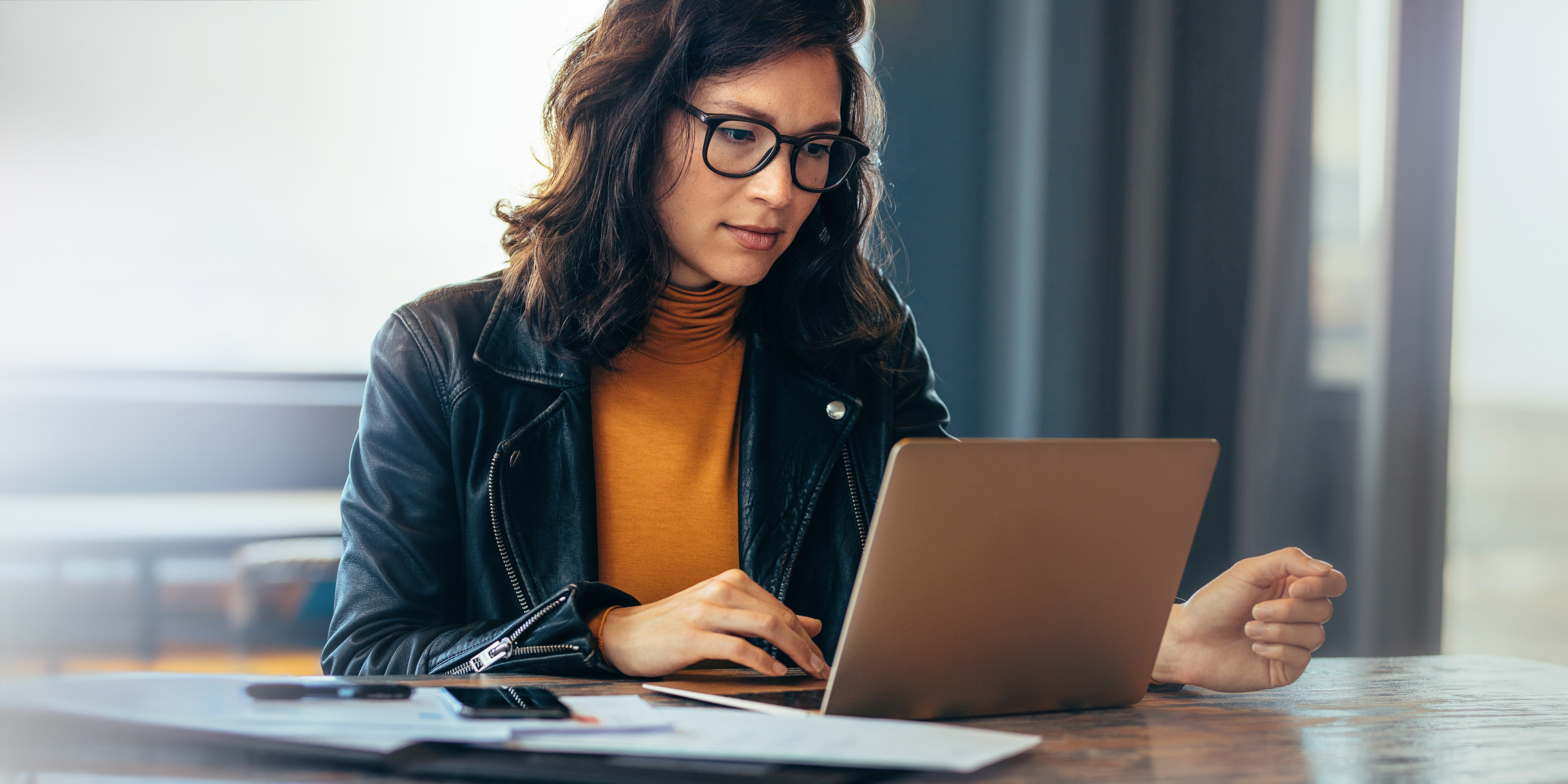 A young woman with dark wavy hair and black-framed glasses, wearing an orange turtleneck and black leather jacket, focused on her laptop at a wooden desk with papers and a phone nearby, in a softly lit office setting.