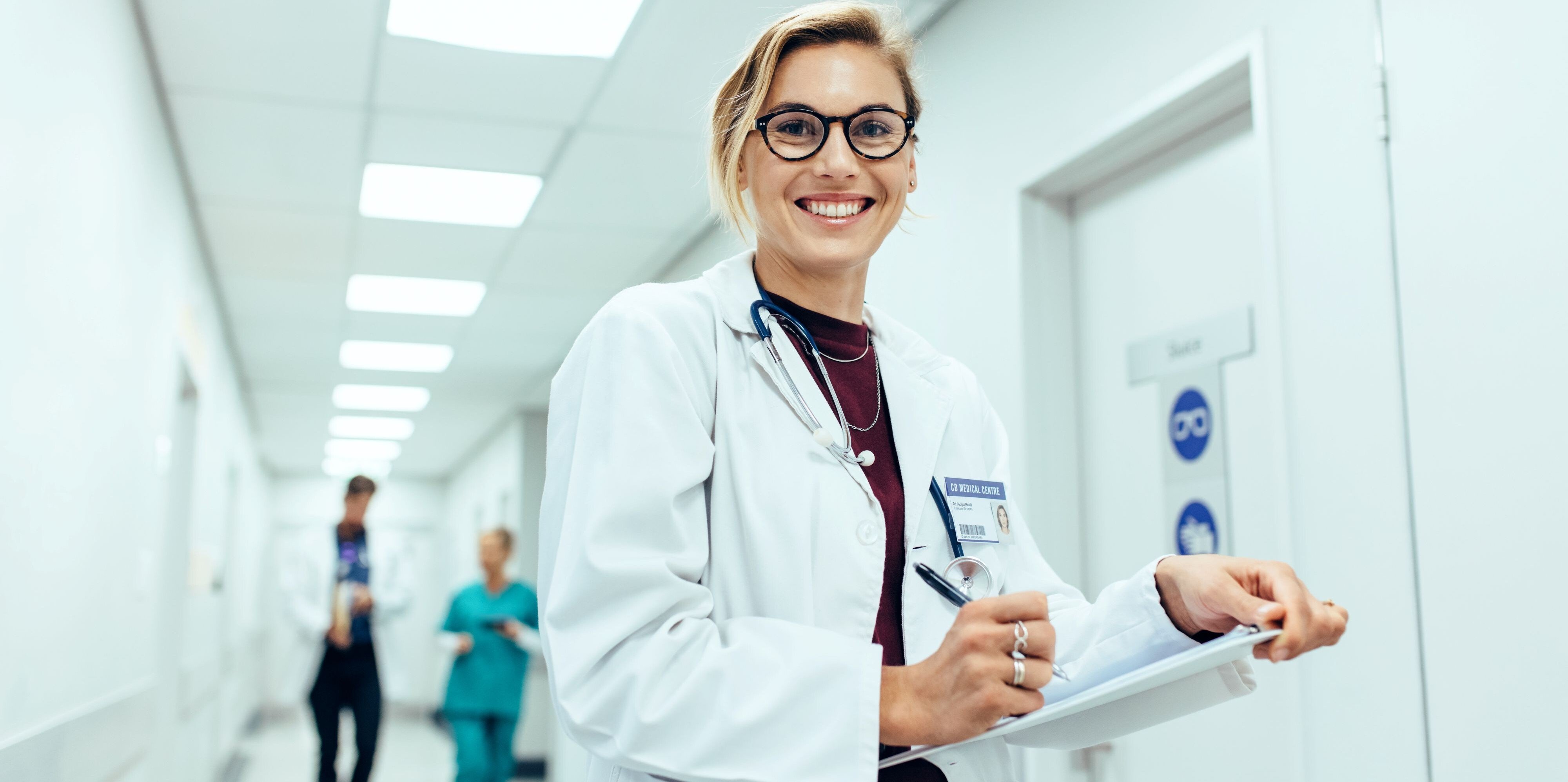 A smiling female physician wearing glasses and a white lab coat with a stethoscope around her neck, standing in a hospital hallway while writing on a clipboard, with other medical staff blurred in the background.