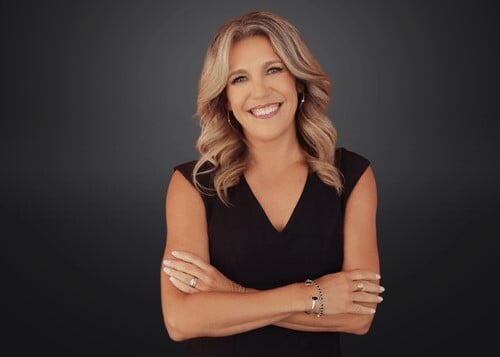 Image of Kelly Rakowski, CEO of America Healthcare Staffing Group and Trio Workforce Solutions. Kelly is standing in front of a dark background with her arms crossed and smiling. 