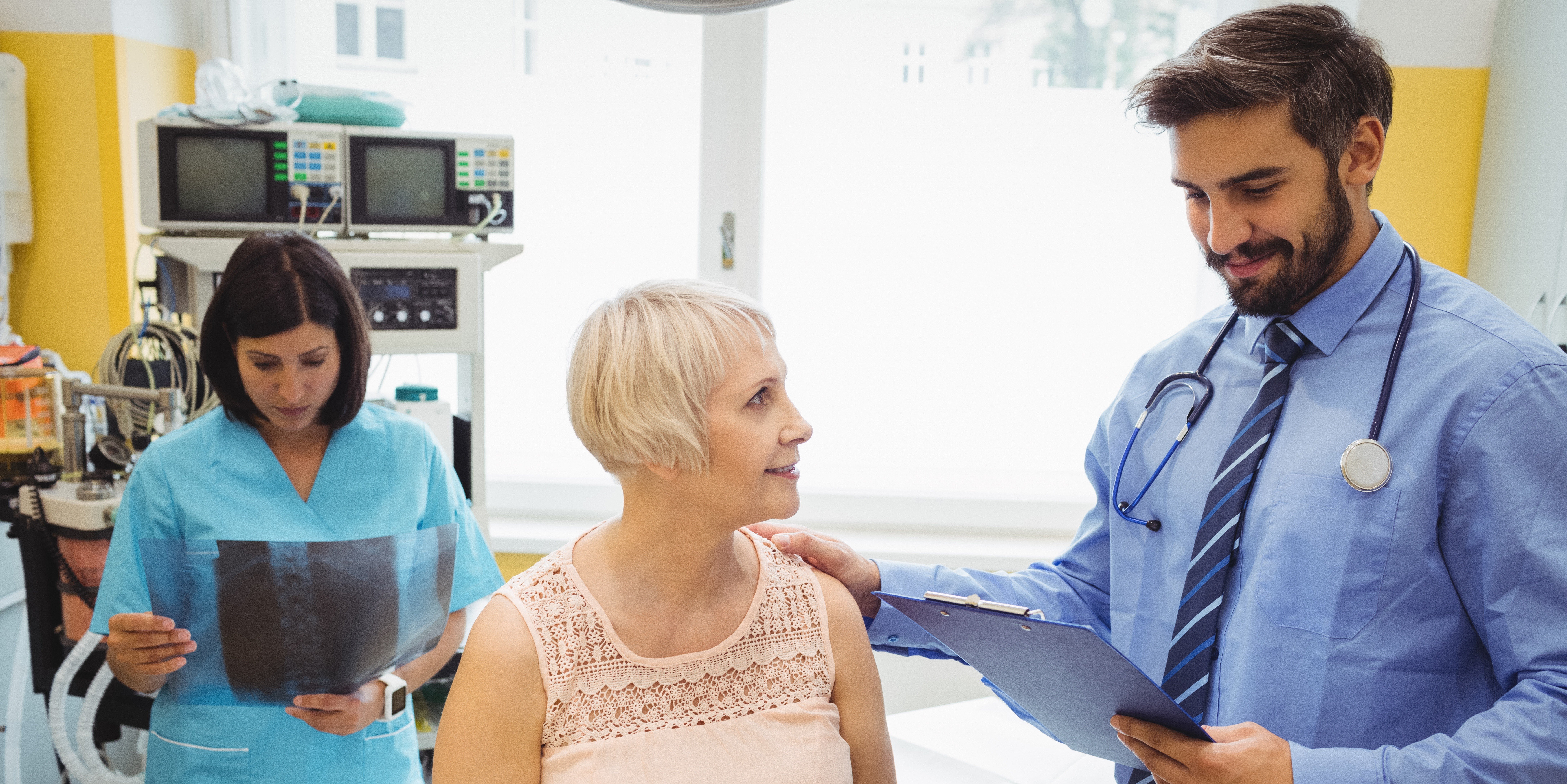 A doctor with a stethoscope reviews paperwork while speaking reassuringly with an older female patient in a hospital exam room. In the background, a nurse in scrubs examines an X-ray beside medical equipment.