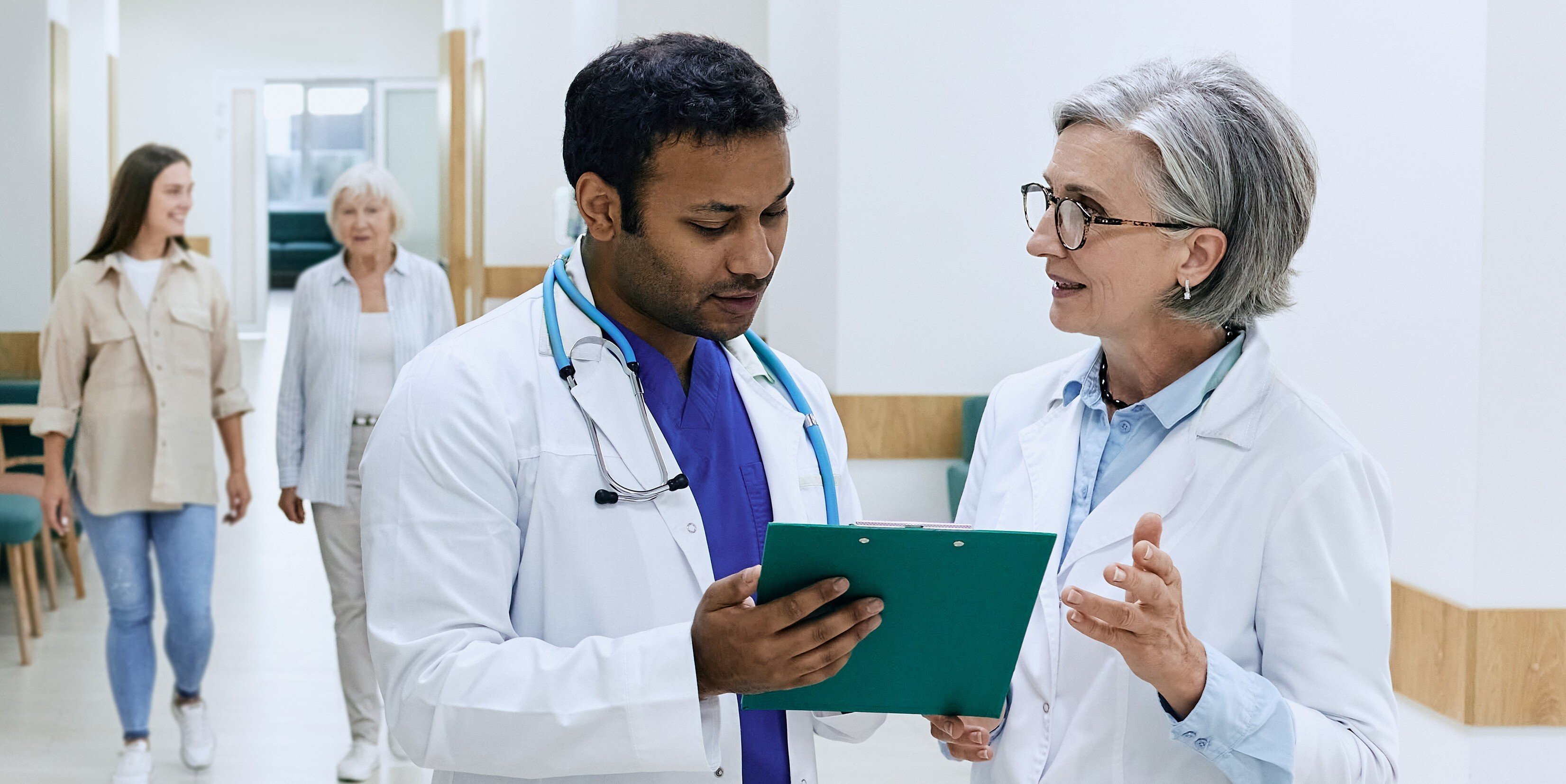 Two healthcare professionals review patient information on a clipboard while walking through a clinic hallway, with patients in the background.