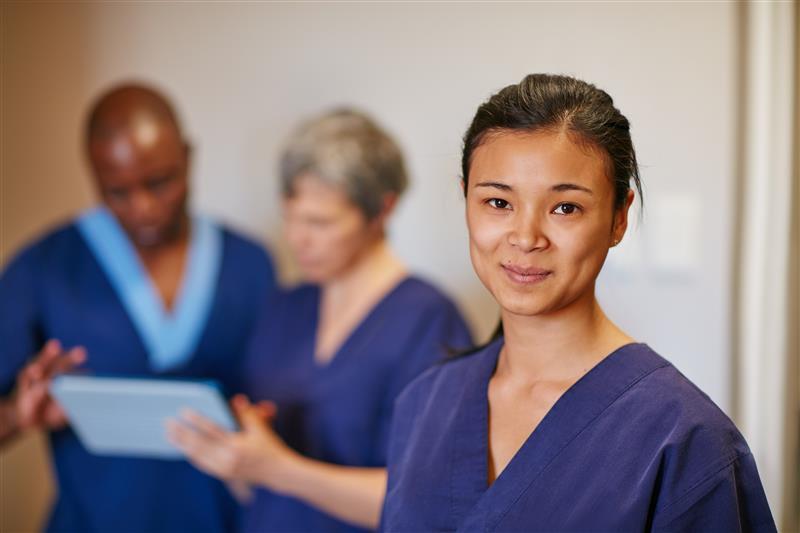 A nurse in blue scrubs is standing and smiling at the camera. Behind her right shoulder is another nurse. She is holding a tablet and showing it to a male nurse.