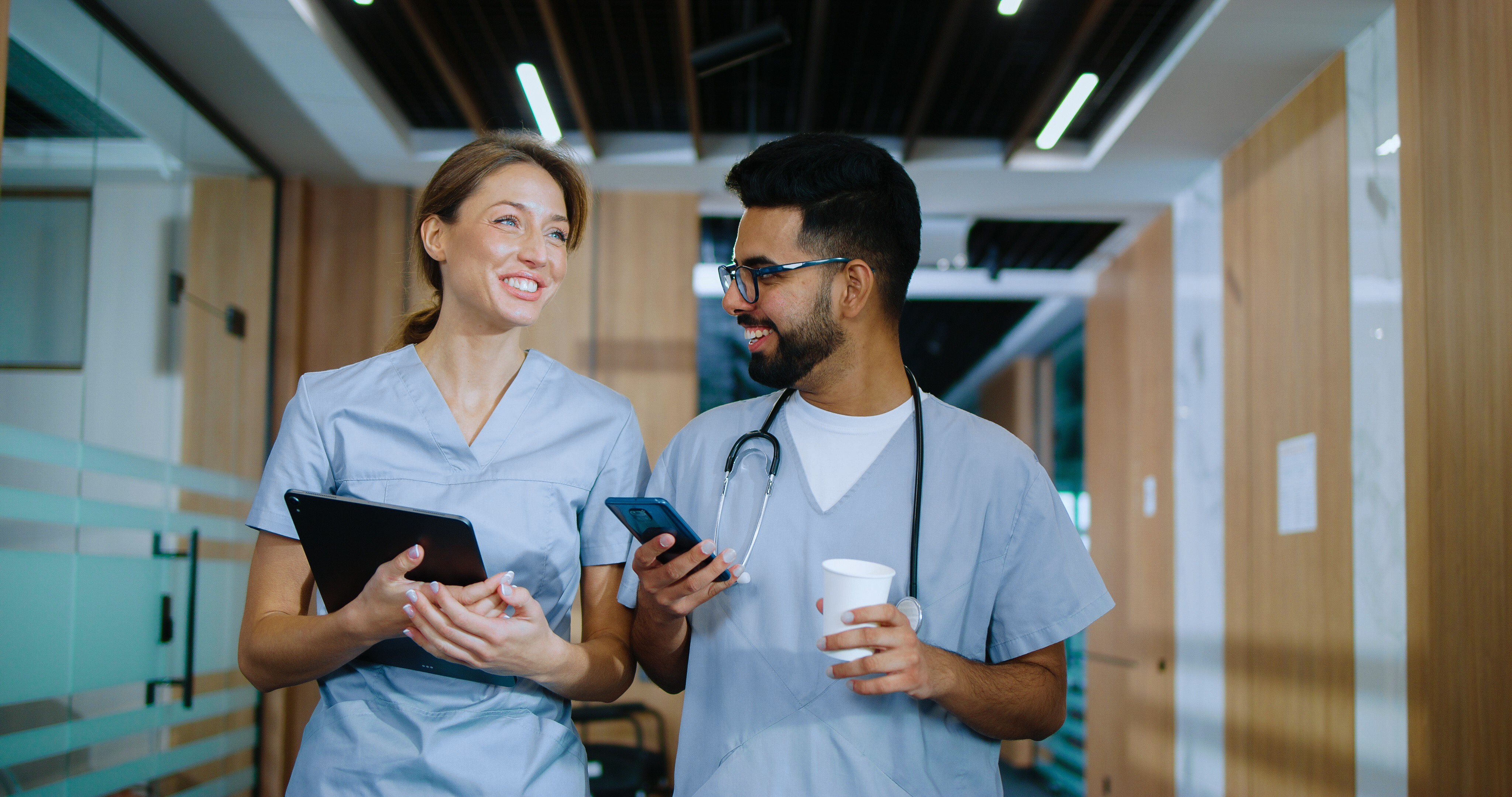 Two healthcare professionals in scrubs walk together down a modern hospital corridor, smiling and talking. One holds a tablet, while the other carries a smartphone, a coffee cup, and wears a stethoscope, suggesting a collaborative and positive clinical work environment.