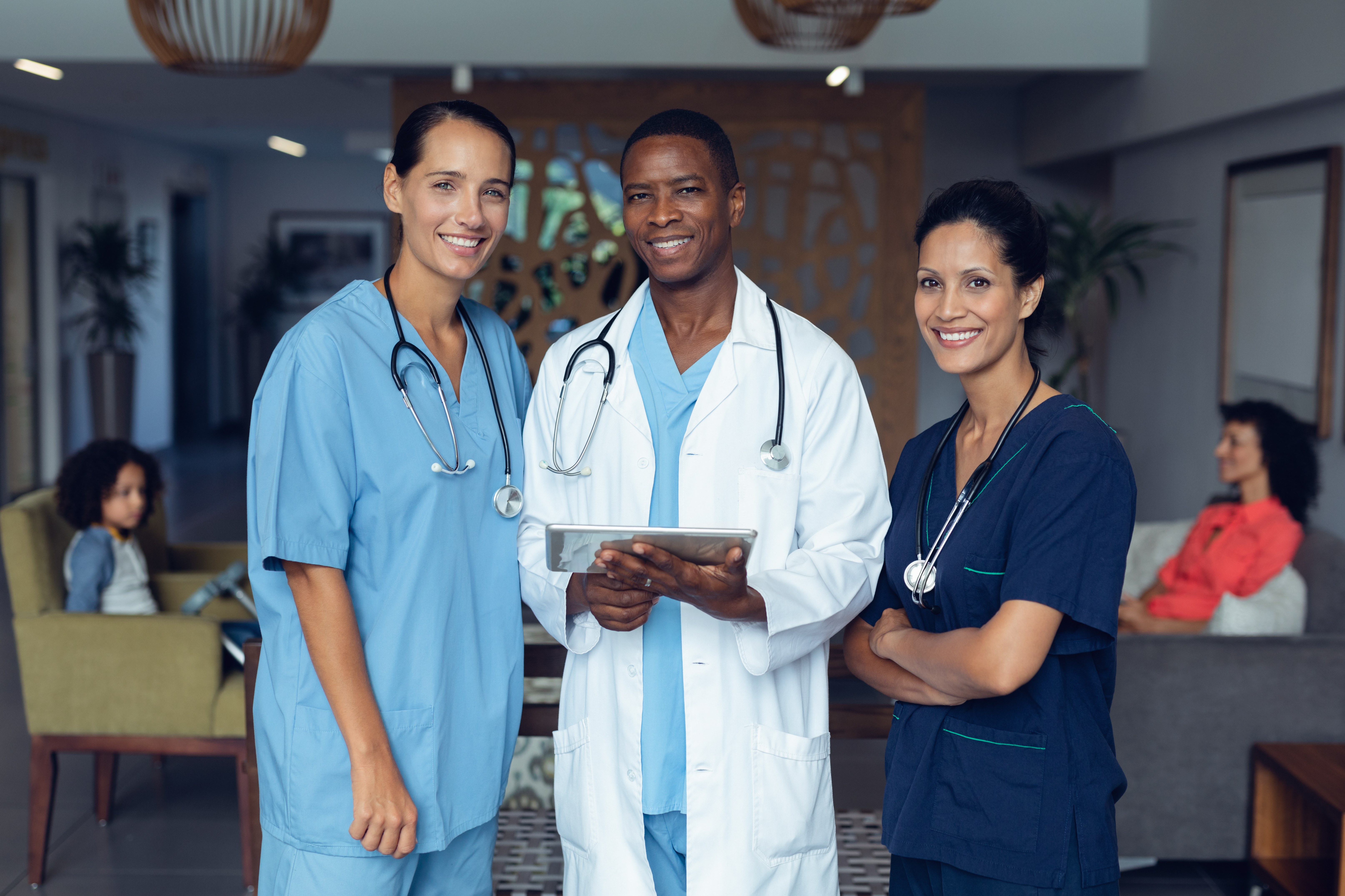 Three healthcare professionals, two nurses and a physician, standing together in a clinical setting, smiling and reviewing information on a tablet, with patients seated in the background.