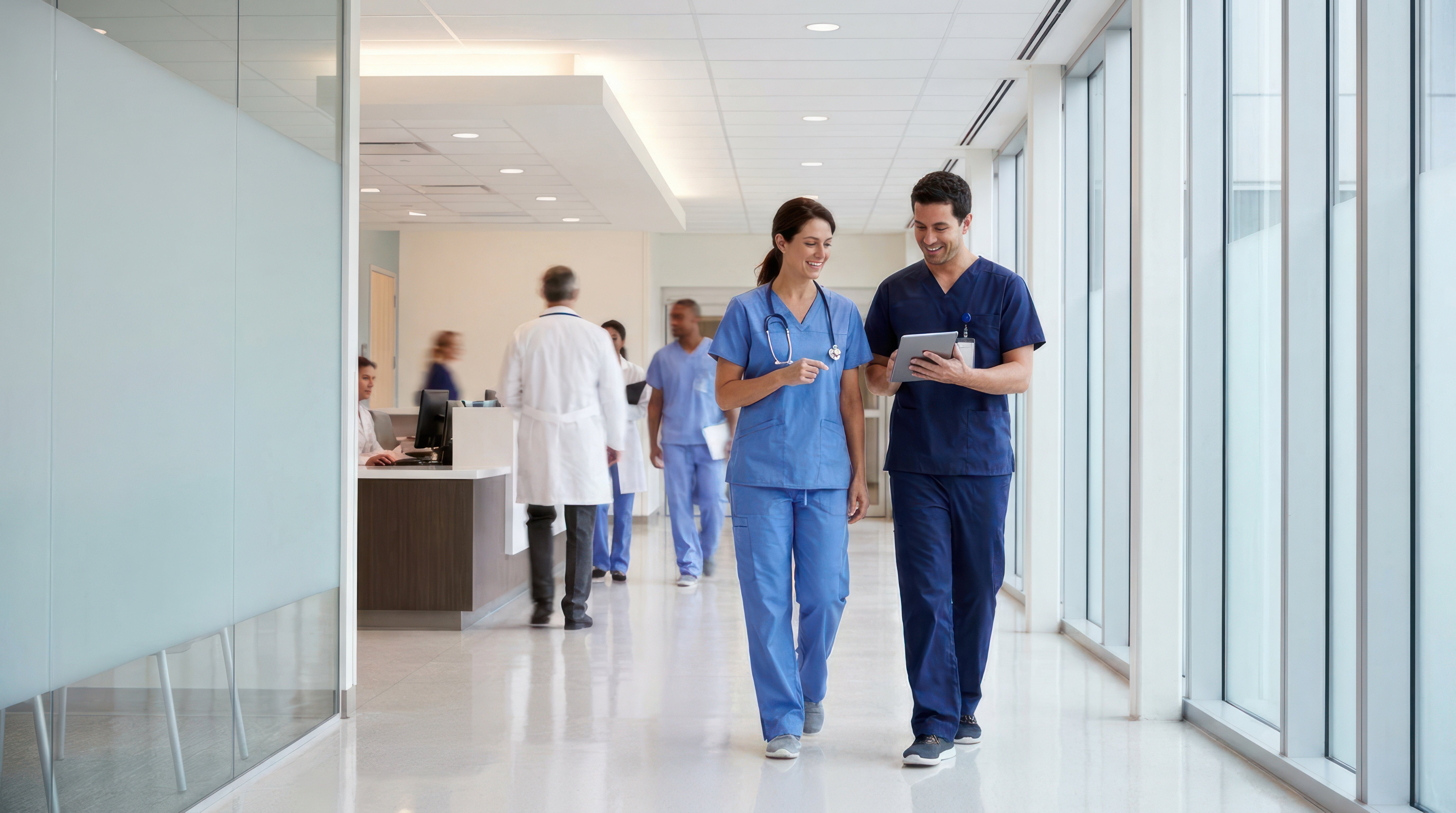 Two clinical staff members in scrubs walk through a hospital corridor reviewing a tablet, with additional healthcare workers visible at a nursing station in the background.