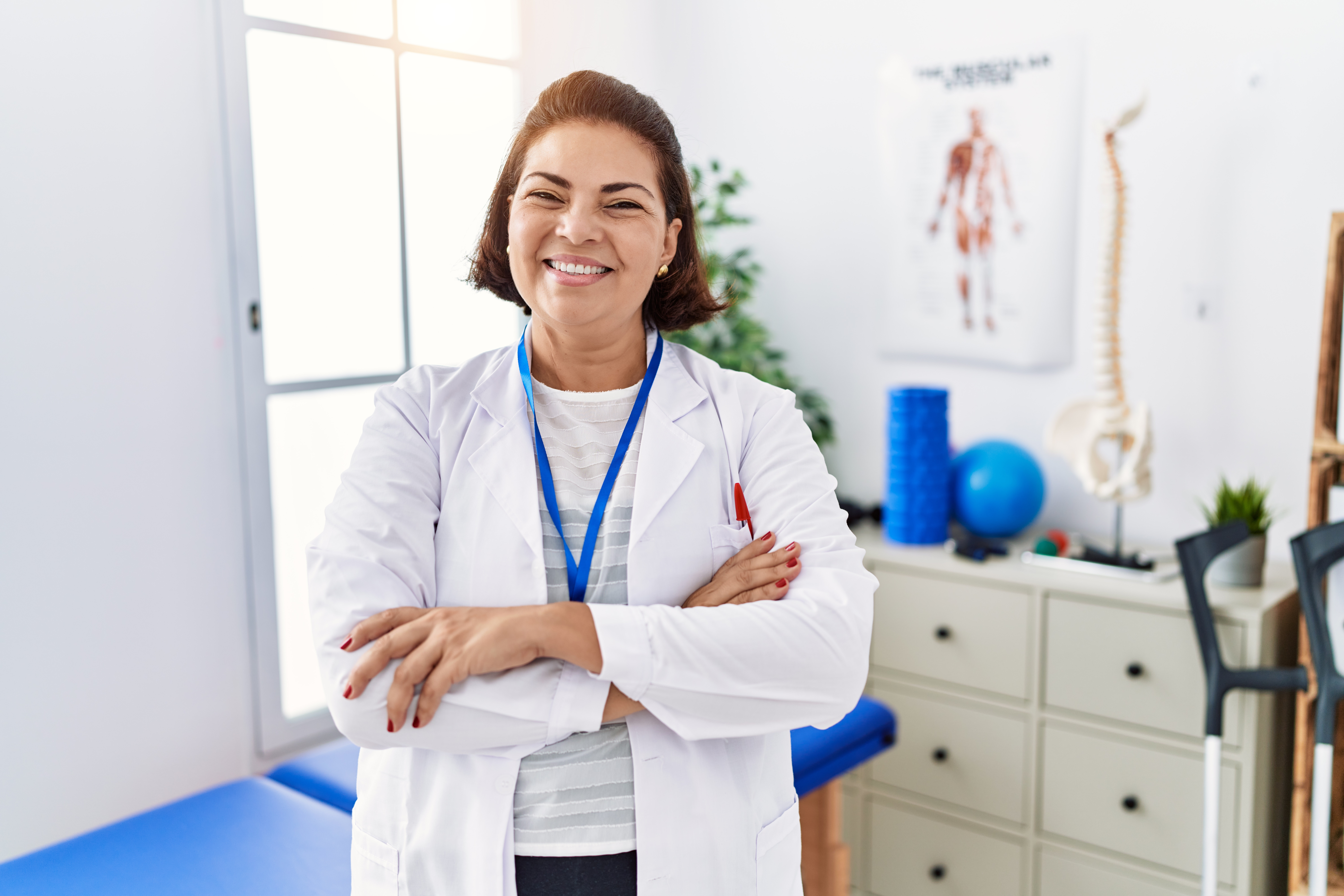 A female physicial therapist stands in the middle of the room. She is smiling, with her arms crossed. Behind her is a therapy bed. It backs up a set of drawers and window. On the wall there is a poster of the human skeleton and muscles. 