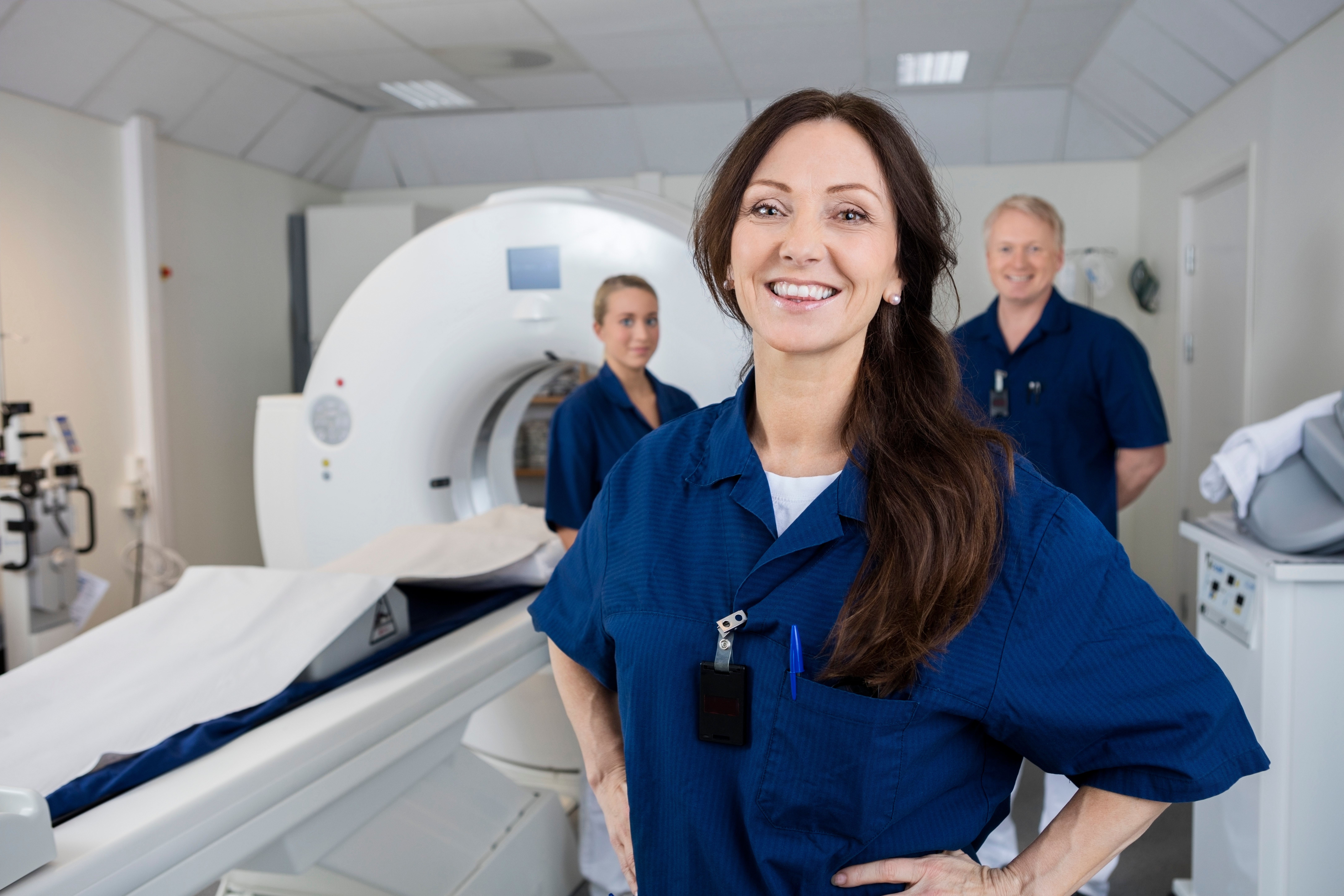 Three rad techs stand in white room in front of an MRI machine. The large white cylinder-shaped machine is on the left with various other machines and devices on the left and right side of the room. Front right is a woman with long dark hair, in blue scrubs, smiling with her hands on her hips. Over her left shoulder is an older man with blonde hair, also in blue scrubs, smiling with his hands tucked behind his back. To his right, and over the first woman's right shoulder, is another woman. This second woman has her sandy-blonde hair pulled up, possibly into a ponytail. She is also in blue scrubs and is staring straight ahead with a slight smile.