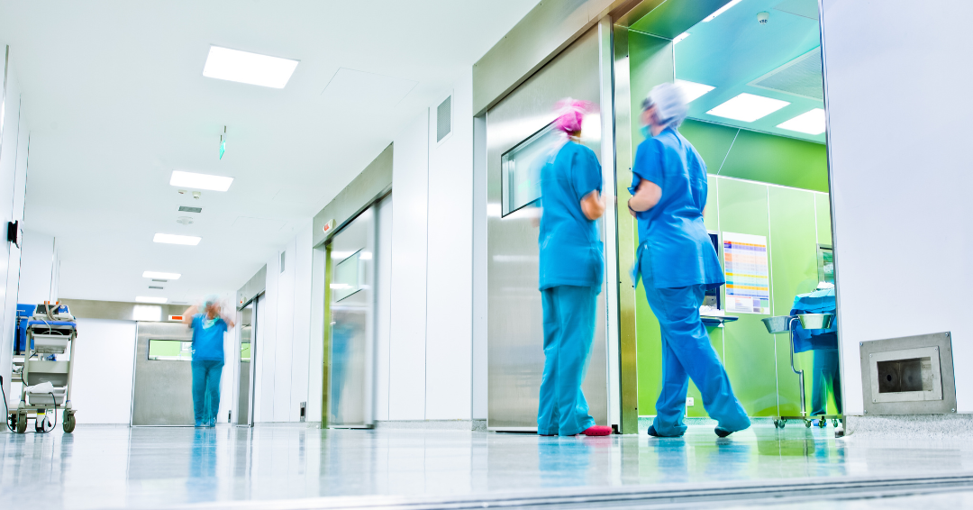 Hospital corridor with medical staff in blue surgical scrubs and hair covers walking past operating room doors, with equipment carts visible and bright, clean clinical lighting throughout the hallway.