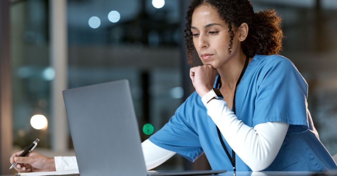Healthcare professional in blue scrubs reviews information on a laptop while taking notes at a desk in a clinical or hospital setting.