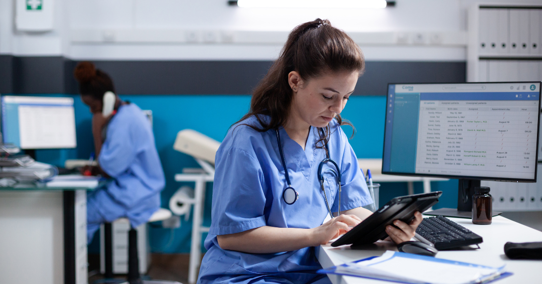 A nurse in blue scrubs sits at a desk in a clinical office, using a tablet to review patient information. A computer monitor displaying a medical schedule is beside her, while another healthcare worker in scrubs speaks on the phone in the background.