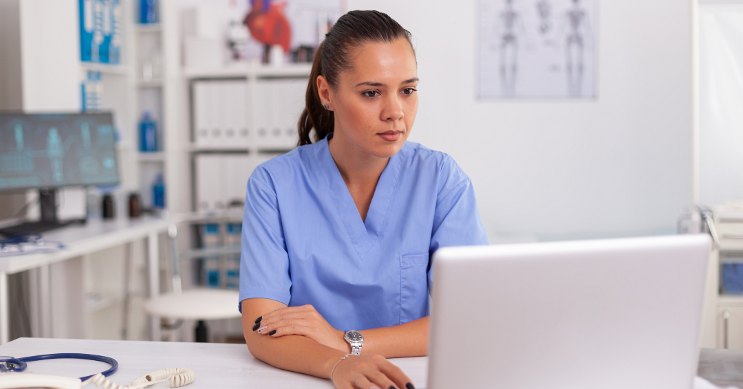 Healthcare professional in blue scrubs seated at a desk, focused on working on a laptop in a clinical office setting.