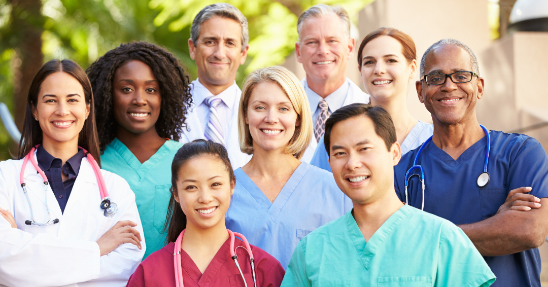 Diverse group of healthcare professionals, including doctors and nurses, standing together outdoors and smiling, representing a collaborative and inclusive healthcare workforce.