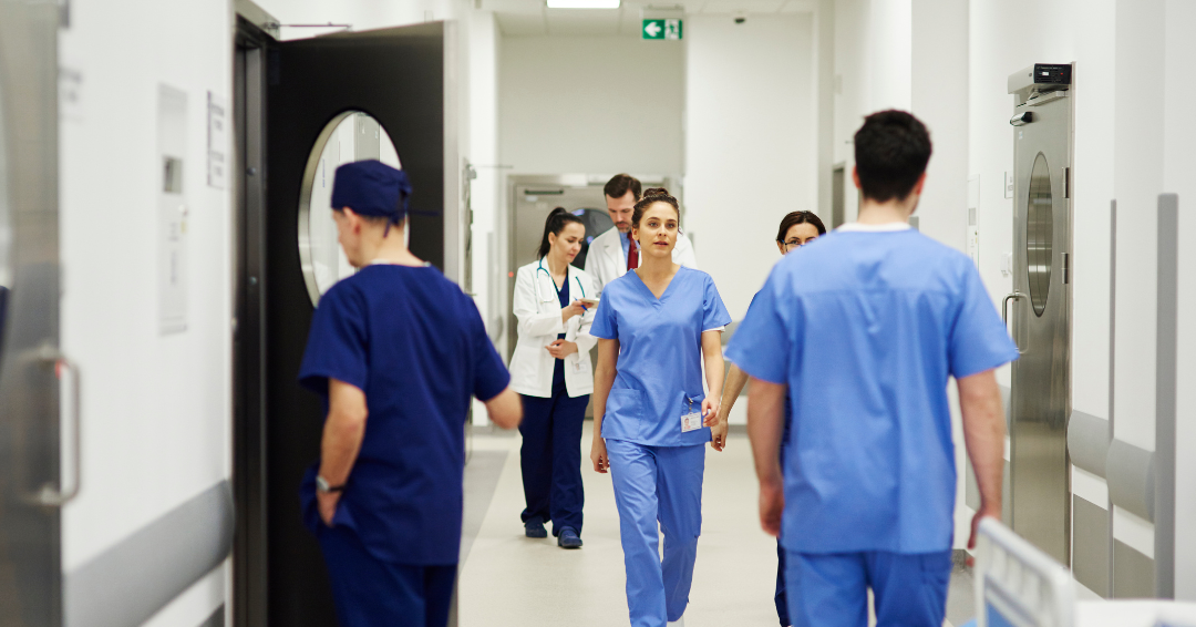 Healthcare professionals in blue scrubs walk through a bright hospital corridor, with doctors and nurses moving between patient care areas and clinical equipment visible along the hallway.
