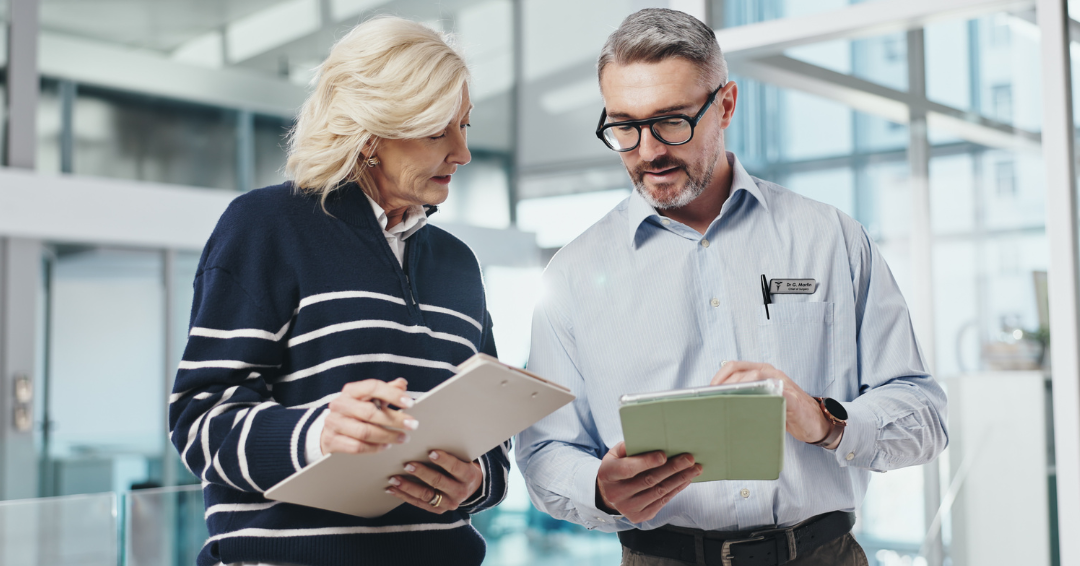 Two healthcare professionals reviewing documents on tablet devices together in a modern hospital or clinical office setting, discussing staffing or operational data.